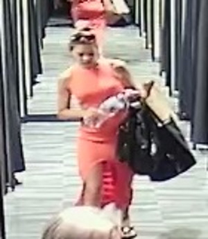 Zdjęcie przedstawia dwie kobietę w sklepie odzieżowym. Ma około 30 lat, około 170 cm wzrostu, szczupła budowa ciała, upięte blond włosy, ubrana w pomarańczową sukienkę, na ramieniu ma dwie czarne torebki, w rękach trzyma jakiś przedmiot. Po prawej stronie zdjęcia widać zasłony pomieszczenia przebieralni.