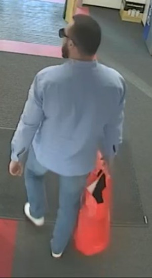 Zdjęcie przedstawia stojącego tyłem do kamery monitoringu mężczyznę, ma on około 180 cm wzrostu, ciemne krótkie włosy, ciemna broda, na twarzy ma założone czarne okulary, ubrany jest w niebieską koszulę, niebieskie spodnie, białe buty, w prawej dłoni trzyma czerwoną torbę zakupową.