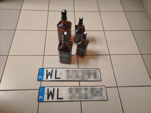 Zdjęcie przedstawia cztery butelki markowego alkoholu stojące na podłodze korytarza, a przed nimi leżą dwie tablice rejestracyjne o wyróżniku WL, reszta treści jest zamazana.