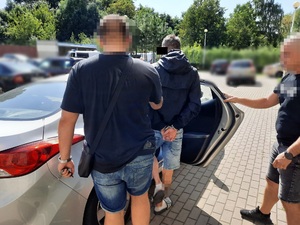 Zdjęcie przedstawia policjanta ubranego w ciemną koszulkę na krótki rękaw i granatowe spodenki jeansowe do kolan, który umieszcza zatrzymanego mężczyznę w zaparkowanym samochodzie. Twarz policjanta jest zasłonięta komputerowym filtrem. Stoi tyłem do obiektywu aparatu. Zatrzymany mężczyzna ubrany jest w czarną ortalionową kurtkę, krótkie jeansowe spodenki w kolorze niebieskim, białe skarpetki i czarne klapki. Część twarzy zasłania mu czarny prostokąt. Mężczyzna na rękach trzymanych z tyłu ma założone kajdanki. Stoi tyłem do obiektywu aparatu. Po jego prawej stronie stoi inny nieumundurowany policjant, który przytrzymuje otwarte drzwi pojazdu umożliwiając zatrzymanemu wejście do środka. Stoi on lewym profilem do obiektywu aparatu. Jego twarz jest zasłonięta komputerowym filtrem. W tle widać inne zaparkowane na dziedzińcu pojazdy.