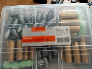 Zdjęcie przedstawia plastikowe przezroczyste pudełko, w którym widać naboje do broni w kolorze ciemnoszarym i złotym, ułożone w osobnych przegródkach.