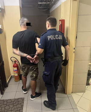 Zdjęcie przedstawia umundurowanego policjanta, który ma na rękach czarne rękawiczki ochronne. Po jego lewej stronie stoi zatrzymany mężczyzna. Ma on siwe włosy, część jego twarzy zasłania czarny prostokąt. Mężczyzna ten ubrany jest w czarną koszulkę na krótki rękaw, ciemnozielone spodenki we wzory o długości do kolan i czarne buty sportowe. Na rękach trzymanych z tyłu ma założone kajdanki. Obaj stoją tyłem do obiektywu aparatu.