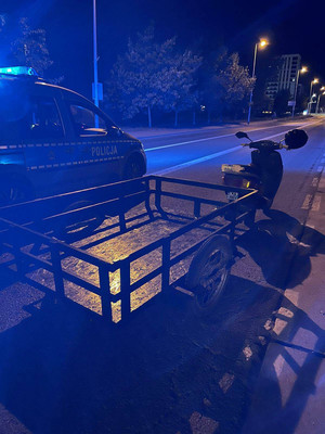Zdjęcie przedstawia skuter, do którego dołączona jest przyczepka. Obok niego, po lewej stronie zdjęcia jest zaparkowany radiowóz policyjny emitujący niebieskie sygnały świetlne. Zdjęcie jest wykonane porą wieczorową.