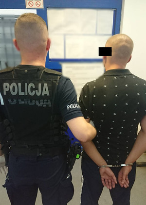 Zdjęcie przedstawia dwóch tyłem stojących do obiektywu aparatu mężczyzn. Po lewej stronie zdjęcia widać umundurowanego policjanta, który przytrzymuje za ramię stojącego po jego prawej stronie zatrzymanego mężczyznę. Mężczyzna ten jest ubrany w czarną koszulkę na krótki rękaw w białe wzory i ciemne długie spodnie. Na rękach trzymanych z tyłu ma założone kajdanki. Część jego twarzy zasłania czarny prostokąt. W tle widać tablicę z białymi kartkami.