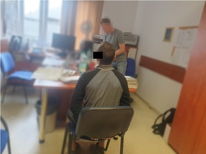 Zdjęcie przedstawia policjanta ubranego w szarą koszulkę na krótki rękaw, który stoi przed biurkiem w pokoju służbowym. Twarz policjanta jest zamazana komputerowym filtrem. Zatrzymany mężczyzna jest ubrany w szarą bluzę z czarnymi długimi rękawami. Siedzi na krześle tyłem do drzwi i obiektywu aparatu. Na rękach trzymanych z tyłu ma założone kajdanki. Część jego twarzy jest zasłonięta czarnym prostokątem.