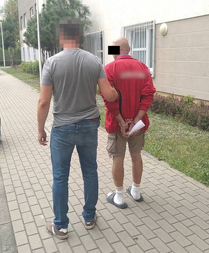 Zdjęcie przedstawia policjanta prowadzącego po dziedzińcu zatrzymanego mężczyznę. Policjant jest bez munduru. Ma na sobie szarą koszulkę na krótki rękaw i niebieskie spodnie jeansowe. Ma twarz zasłoniętą filtrem komputerowym. Po swojej prawej stronie prowadzi zatrzymanego mężczyznę. Mężczyzna ten jest łysy, część jego twarzy zasłania czarny prostokąt. Ubrany jest w czerwoną bluzę na długi rękaw, krótkie beżowe spodenki, białe skarpety i czarne klapki. Na rekach trzymanych z tyłu ma założone kajdanki. Obaj znajdują się tyłem do obiektywu aparatu. Idą wzdłuż budynku komendy z okratowanymi oknami.