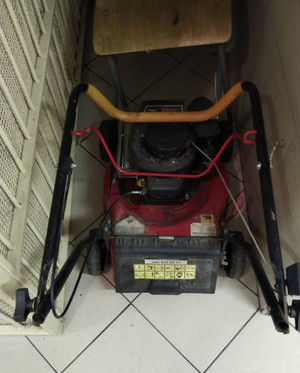 Zdjęcie przedstawia stojącą na szarej posadzce czerwono-czarną kosiarkę.