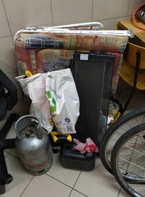 Zdjęcie przedstawia ułożone pod ścianą przedmioty: łóżko polowe z kolorowym materacem, szarą butlę gazową, czarny pojemnik na paliwo i fragment kół rowerowych.