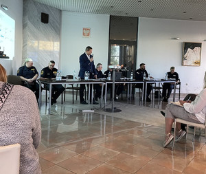 Zdjęcie przedstawia przedstawicieli policji, straży miejskiej i prokuratury, którzy siedzą w długim stole przed uczestnikami spotkania. Mężczyzna w garniturze stoi trzymając przy ustach mikrofon.