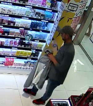 Zdjęcie przedstawia mężczyznę stojącego wewnątrz drogerii. Jest on skierowany lewym profilem do kamery. Ma on około 35-40 lat, 175 cm wzrostu, ubrany jest w ciemną koszulę, niebieskie spodnie jeansowe, czapkę z daszkiem, czerwone sportowe buty. Przez prawą rękę ma przewieszoną kurtkę. W tle widać półki z asortymentem sklepowym.