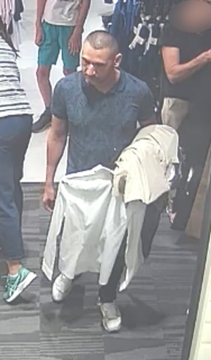 Zdjęcie przedstawia mężczyznę w wieku około 35-40 lat, szczupłej budowy ciała, włosy ostrzyżone po bokach i z tyłu, ubrany w ciemnozieloną koszulkę, ciemne spodnie, białe buty. Na lewej ręce ma przewieszone ubrania w jasnym kolorze, w lewej dłoni trzyma wieszak z jasną koszulą. Mężczyzna jest skierowany przodem do kamery. W tle widać fragmenty sylwetek innych osób.