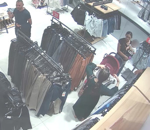 Zdjęcie przedstawia trzy osoby stojące pomiędzy wieszakami w sklepie. Mężczyzna jest  w wieku około 35-40 lat, szczupłej budowy ciała, ma włosy ostrzyżone po bokach i z tyłu, ubrany jest w ciemnozieloną koszulkę, ciemne spodnie. Na lewej ręce ma przewieszone ubrania. Mężczyzna jest skierowany przodem do kamery. Na tym samym zdjęciu jest kobieta w wieku około 30-35 lat, o ciemniejszej karnacji, ubrana w czarną koszulkę na krótki rękaw, ciemnozieloną spódnicę, trzyma ona na rekach małe dziecko. Przed nią stoi wózek dziecięcy w kolorze bordowym. W dole zdjęcia jest kobieta w wieku około 30-35 lat, o ciemniejszej karnacji, ubrana w długą sukienkę koloru zielonego, czarne klapki. Kobieta stoi tyłem do kamery, trzyma w dłoniach jakieś ubranie.