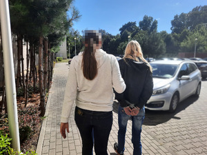 Zdjęcie przedstawia policjantkę po cywilnemu, w jasnej bluzie i ciemnych spodniach jeansowych, która prowadza po dziedzińcu zatrzymaną kobietę. Twarz policjantki jest zasłonięta komputerowym filtrem. Zatrzymana kobieta ma długie blond włosy, czarną kurtkę i ciemne spodnie jeansowe. Na rekach trzymanych z tyłu ma założone kajdanki. Obie są tyłem do obiektywu aparatu. W tle widać zaparkowane pojazdy.