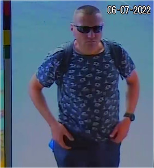 Zdjęcie przedstawia mężczyznę w wieku około 40 lat, 185 cm wzrostu, masywnej budowy ciała; ubrany jest w koszulkę typu t-shirt koloru ciemnego z białymi znaczkami, na twarzy miał czarne okulary przeciwsłoneczne. Na lewej ręce miał czarny pasek od zegarka. Stoi przodem do kamery.