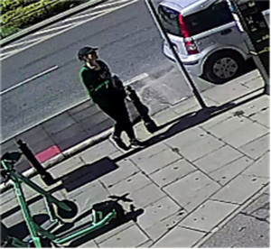 Zdjęcie przedstawia kobietę stojącą na chodniku. Kobieta jest w wieku około 20-30 lat, szczupłej budowy ciała; ubrana jest w zieloną bluzę, czarne spodnie legginsy, czarne buty sportowe z białą podeszwą, czarną czapkę z daszkiem. W tle widać zaparkowane pojazdy.