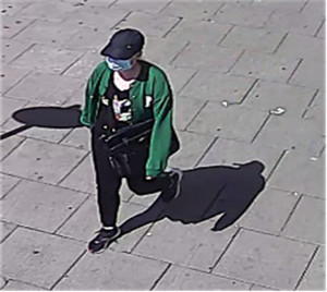 Zdjęcie przedstawia kobietę idącą chodnikiem. Kobieta jest w wieku około 20-30 lat, szczupłej budowy ciała; ubrana jest w zieloną bluzę, czarne spodnie legginsy, czarne buty sportowe z białą podeszwą, czarną czapkę z daszkiem. Na twarzy ma maseczkę ochronną. W tle widać zaparkowane pojazdy.