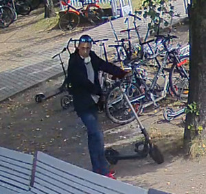 Zdjęcie przedstawia mężczyznę w wieku około 30 lat, około 170-175 cm wzrostu. Ubrany jest w czarną kurtkę, niebieskie spodnie, czerwone buty sportowe, prowadzi hulajnogę. W tle widać pozostawione na trawniku rowery i hulajnogi.