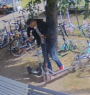 Zdjęcie przedstawia mężczyznę ubranego w czarną kurtkę, niebieskie spodnie, czerwone buty sportowe, który pochyla się nad hulajnogą. W tle widać pozostawione na trawniku rowery i hulajnogi.