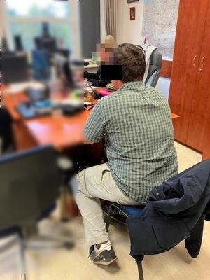 Zdjęcie przedstawia policjantkę, która przesłuchuje zatrzymanego mężczyznę. Twarz policjantki jest zasłonięta. Część twarzy mężczyzny również zasłania czarny prostokąt. Mężczyzna ten siedzi przy biurku, tyłem do drzwi wejściowych do pokoju. Jest ubrany w jasną koszulę w kratę na krótki rękaw, długie jasne spodnie i jasne buty sportowe.