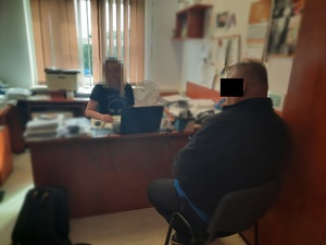 Zdjęcie przedstawia policjantkę, która siedzi przy biurku, jej twarz jest zasłonięta. Naprzeciwko niej, przy tym samym biurku, tyłem do drzwi siedzi zatrzymany mężczyzna. Jest on ubrany na ciemno. Część jego twarzy zasłania czarny prostokąt.