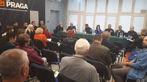 Zdjęcie przedstawia przedstawicieli służb i instytucji, którzy siedzą za długim stołem przed zebranymi w sali mieszkańcami. Mieszkańcy siedzą tyłem do wejścia do sali.