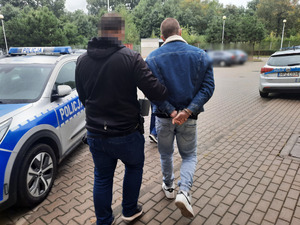 Zdjęcie przedstawia policjanta ubranego w czarną kurtkę i granatowe spodnie jeansowe. Po jego prawej stronie idzie zatrzymany 18-latek. Ma on niebieską kurtkę jeansową i niebieskie spodnie jeansowe. Na rękach trzymanych z tyłu ma założone kajdanki. Część twarzy zasłania mu czarny prostokąt. W tle widać zaparkowany oznakowany radiowóz.