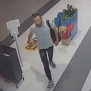 Zdjęcie przedstawia mężczyznę wchodzącego do sklepu. Ma on ciemne włosy, okulary przeciwsłoneczne na głowie, jest ubrany w szarą koszulkę na krótki rękaw, czarne długie spodnie z białymi wstawkami i biało-czarne buty sportowe. Przez lewe ramię ma przewieszoną czarną torbę.