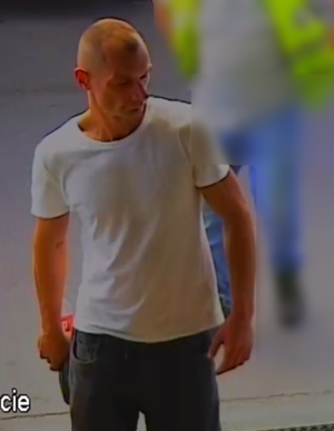 Zdjęcie przedstawia mężczyznę wchodzącego do obiektu. Jest on ustawiony prosto do kamery. Ma krótkie jasne włosy, ubrany jest w białą koszulkę na krótki rękaw i granatowe spodnie.