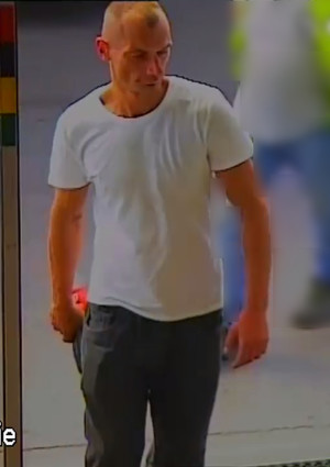 Zdjęcie przedstawia mężczyznę wchodzącego do obiektu. Jest on ustawiony prosto do kamery. Ma krótkie jasne włosy, ubrany jest w białą koszulkę na krótki rękaw i granatowe spodnie.