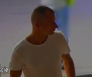 Zdjęcie przedstawia fragment sylwetki mężczyzny wchodzącego do obiektu. Jest on ustawiony prawym profilem do kamery. Ma krótkie jasne włosy, ubrany jest w białą koszulkę na krótki rękaw.