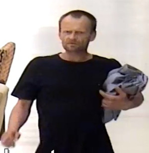 Zdjęcie przedstawia mężczyznę w wieku około 40 lat,ma  około 175 cm wzrostu, krótkie ciemne włosy, widoczny ciemny zarost, ubrany jest w czarną koszulkę na krótki rękaw. Lewą ręką dociska do lewej strony klatki piersiowej jakiś przedmiot. Jest ustawiony na wprost kamery monitoringu.