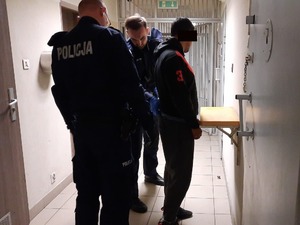 Zdjęcie przedstawia dwóch umundurowanych policjantów, którzy zakładają kajdanki zespolone zatrzymanemu mężczyźnie. Jest on ustawiony przy ścianie. Część jego twarzy zasłania czarny prostokąt.