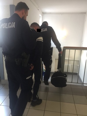 Zdjęcie przedstawia umundurowanych policjantów, którzy prowadzą po korytarzu komisariatu, pomiędzy sobą zatrzymanego mężczyznę. Mężczyzna ten jest ubrany na ciemno, na rekach trzymanych z tyłu ma założone kajdanki. Część twarzy zasłania mu czarny prostokąt. Wszyscy są ustawieni tyłem do obiektywu aparatu.