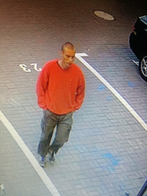 Zdjęcie przedstawia mężczyznę stojącego na miejscu parkingowym. Mężczyzna ten jest w wieku około 25-30 lat, szczupłej budowy ciała, ma krótko ścięte włosy; ubrany jest w czerwoną bluzę i szare spodnie. W tle, po prawej stronie zdjęcia widać fragment zaparkowanego pojazdu w ciemnym kolorze.