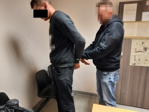 Zdjęcie przedstawia policjanta ubranego w ciemną bluzę i niebieskie spodnie jeansowe, który stoi za zatrzymanym mężczyzną i zakłada mu kajdanki na ręce trzymane z tyłu. Mężczyzna ten jest ubrany na ciemno. Część jego twarzy zasłania czarny prostokąt. Obaj znajdują się w pomieszczeniu dla osób zatrzymanych. Stoją lewym profilem do obiektywu aparatu. Za nimi widać otwarte drzwi do pomieszczenia.