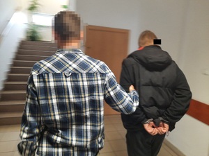 Zdjęcie przedstawia policjanta w koszuli w kratkę, który prowadzi zatrzymanego mężczyznę po korytarzu. Mężczyzna ten ma kajdanki założone na rękach trzymanych z tyłu. Mężczyzna jest ubrany na ciemno. Część jego twarzy zasłania czarny prostokąt. Obaj stoją tyłem do obiektywu aparatu.