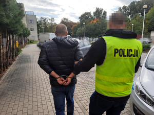 Zdjęcie przedstawia nieumundurowanego policjanta w odblaskowej kamizelce z napisem POLICJA, który prowadzi przez dziedziniec zatrzymanego mężczyznę. Mężczyzna ten jest ubrany w czarną kurtkę, czarne spodnie i buty sportowe. Na rękach trzymanych z tyłu ma założone kajdanki. W tle widać zaparkowane cywilne pojazdy.