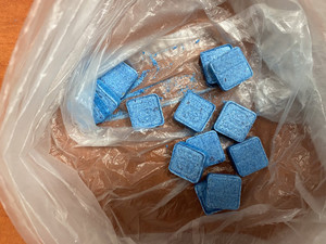 Zdjęcie przedstawia foliową torebkę leżącą na brązowym blacie stołu zawierającą niebieskie tabletki.