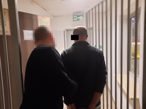 Zdjęcie przedstawia policjanta ubranego na ciemno, który wyprowadza z pomieszczenia zatrzymanego mężczyznę. Obaj są tyłem do obiektywu aparatu. Głowa policjanta jest zasłonięta komputerowym filtrem. Zatrzymany mężczyzna znajduje się po prawej stronie zdjęcia.Na wysokości jego twarzy znajduje się czarny prostokąt. Mężczyzna jest ubrany na ciemno, ma kajdanki założone na rękach trzymanych z tyłu.