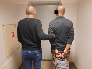 Zdjęcie przedstawia policjanta ubranego na ciemno, który wyprowadza z pomieszczenia zatrzymanego mężczyznę. Obaj są tyłem do obiektywu aparatu. Głowa policjanta jest zasłonięta komputerowym filtrem. Zatrzymany mężczyzna znajduje się po prawej stronie zdjęcia.Na wysokości jego twarzy znajduje się czarny prostokąt. Mężczyzna jest ubrany na ciemno, ma kajdanki założone na rękach trzymanych z tyłu. Trzyma w nich plecak.