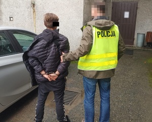 Zdjęcie przedstawia dwie osoby stojące przed budynkiem komisariatu. Po prawej stronie zdjęcia widać policjanta ubranego w odblaskową kamizelkę z napisem POLICJA na plecach. Twarz tego policjanta jest zasłonięta. Przytrzymuje on za przedramię młodego mężczyznę ubranego w ciemną kurtkę i ciemne spodnie, który ma kajdanki założone na rękach trzymanych z tyłu. Część twarzy zasłania mu czarny prostokąt. Obaj stoją tyłem do obiektywu aparatu.
