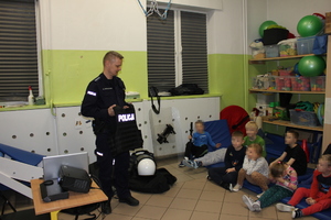 zdjęcie przedstawia umundurowanego policjanta, kry stoi przed grupą dzieci siedzących na podłodze.