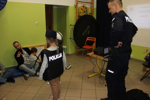 zdjęcie przedstawia umundurowanego policjanta, który stoi za dziewczynką ubraną w elementy umundurowania policyjnego.