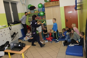 zdjęcie przedstawia umundurowaną policjantkę, która wręcza chłopcu opaskę odblaskową. Inne dzieci siedzą na podłodze z opiekunami.