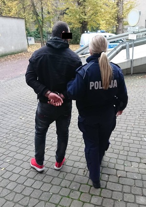 Umundurowana policjantka prowadzi zatrzymanego mężczyznę. Mężczyzna ma kajdanki na rekach trzymanych z tyłu. Część jego twarzy zasłania czarny prostokąt.