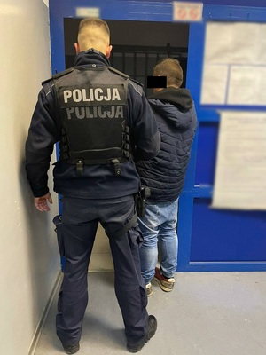 Zdjęcie przedstawia umundurowanego policjanta w czarnej kamizelce taktycznej, który wyprowadza zatrzymanego z pomieszczenia. Mężczyzna ten stoi po prawej stronie policjanta. Jest ubrany w ciemną kurtkę i niebieskie spodnie jeansowe. Część jego twarz zasłania czarny prostokąt. Obaj stoją tyłem do zdjęcia.