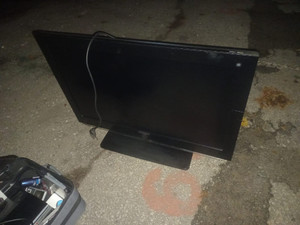 Zdjęcie przedstawia stojący na asfalcie czarny telewizor z płaskim ekranem.