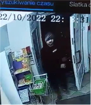 Zdjęcie przedstawia mężczyznę w wieku około 35 lat, normalnej budowy ciała, posiada zarost w postaci wąsów koloru czarnego, nos duży, ubrany jest w czarne dresowe spodnie materiałowe, czarne buty sportowe, czarną kurtkę, czarną bluzkę pod spodem z białą wstawką na wysokości klatki piersiowej, czapkę materiałową z białym logo.