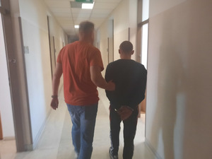 Zdjęcie przedstawia policjanta ubranego w czerwoną koszulkę na krótki rękaw i niebieskie spodnie jeansowe, który prowadzi korytarzem, po swojej prawej stronie zatrzymanego mężczyznę. Twarz policjanta jest zamazana. Mężczyzna jest ubrany na ciemno. Na rękach trzymanych z tyłu ma założone kajdanki.