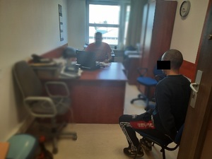 Zdjęcie przedstawia policjanta ubranego w czerwoną koszulkę na krótki rękaw siedzącego za biurkiem, przed otwartym laptopem, przodem do wejścia do pokoju. Jego twarz jest zamazana. Po prawej stronie zdjęcia, lewym profilem do wejścia do pokoju siedzi zatrzymany mężczyzna. Mężczyzna jest ubrany na ciemno. Na rękach trzymanych z tyłu ma założone kajdanki.